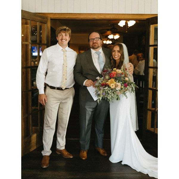 三个人站着摆姿势拍照. 其中一人身穿新娘礼服，手捧鲜花.