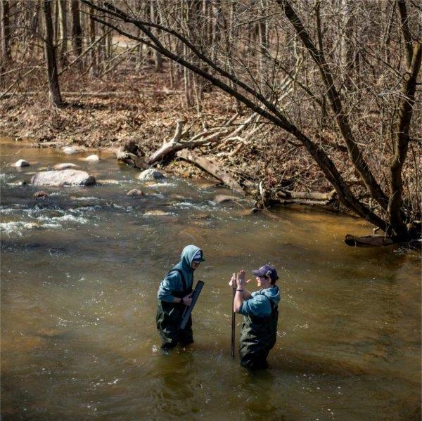 One person gestures to another while both are standing in a stream.